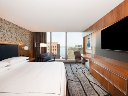 This image shows a modern hotel room with a large bed, wall-mounted TV, desk, chair, and large window offering a city and water view.