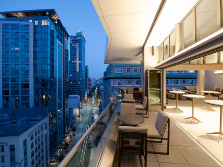 The image shows a modern balcony overlooking an urban cityscape with tall buildings, city lights, and an elegant furnished seating area.