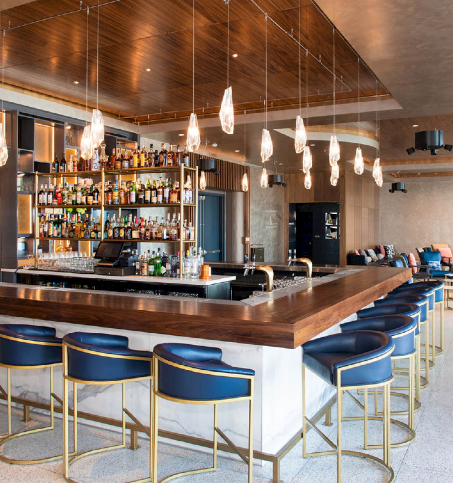 A modern bar with a wooden counter, blue barstools, a fully stocked back shelf, pendant lights, and seating areas in the background.