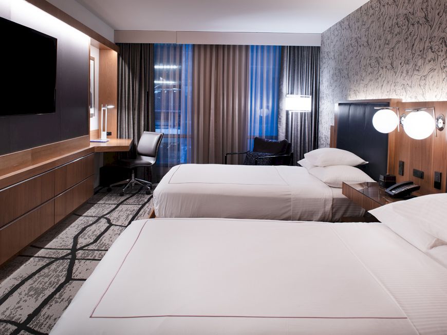 This image shows a modern hotel room with two double beds, a TV, a work desk, and stylish decor. The room has a contemporary design.