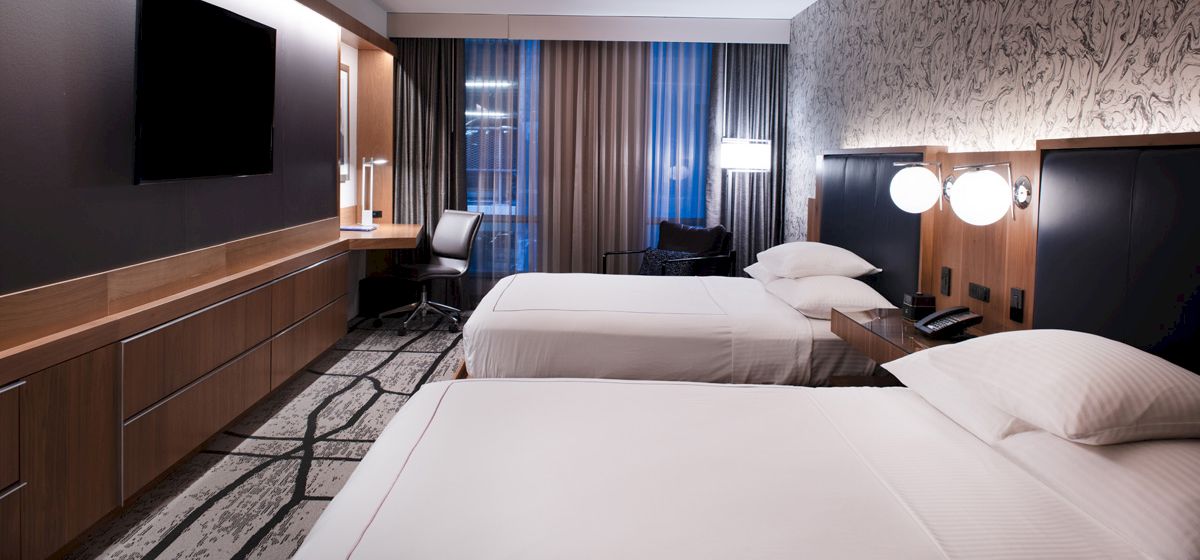 This image shows a modern hotel room with two double beds, a TV, a work desk, and stylish decor. The room has a contemporary design.
