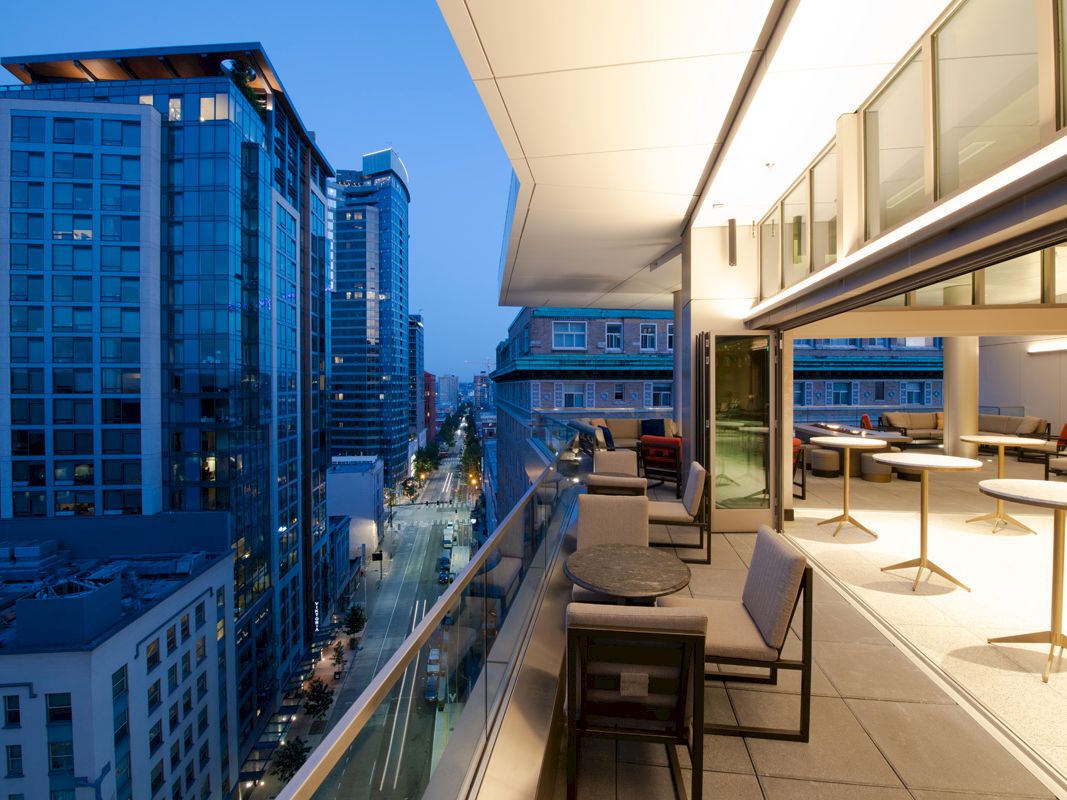 A modern urban balcony with seating overlooks a cityscape filled with high-rise buildings at twilight, with lights illuminating the street below.