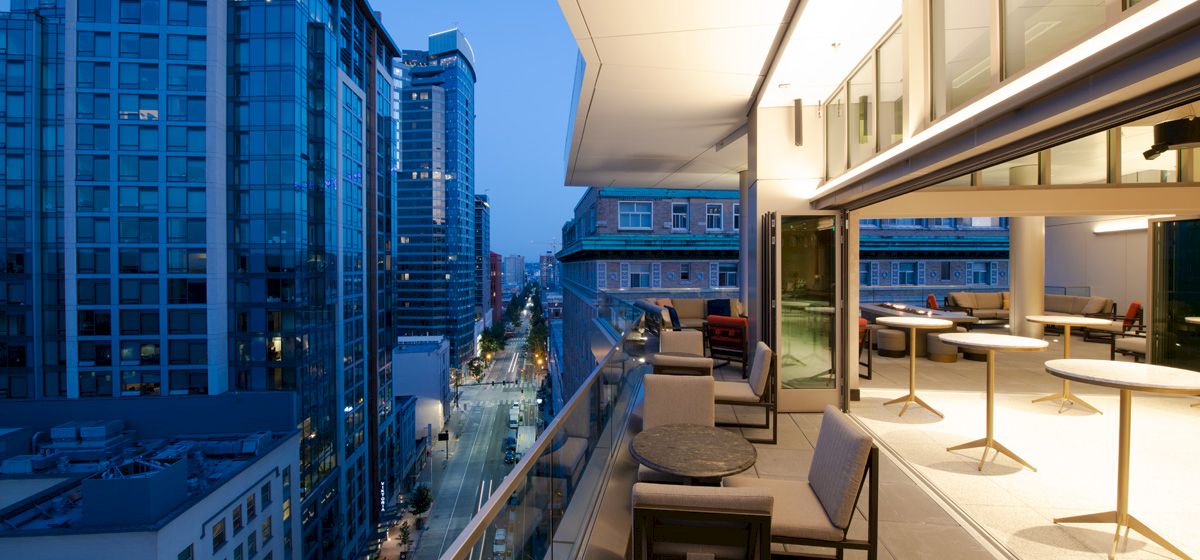 A modern urban balcony with seating overlooks a cityscape filled with high-rise buildings at twilight, with lights illuminating the street below.