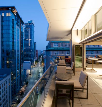 A modern balcony overlooking a city street, with tables, chairs, and buildings illuminated at dusk on both sides.