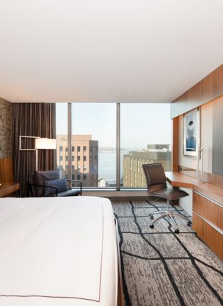 This image shows a modern hotel room with a large bed, desk, chair, flat-screen TV, and a window view of city buildings and water.