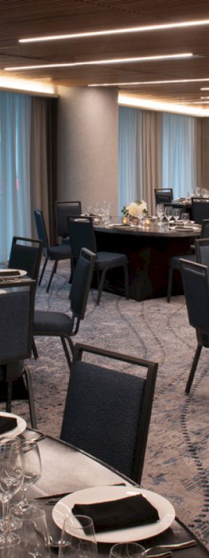 The image shows an elegant, modern banquet hall with round tables set for a formal event. The tables have black tablecloths, folded napkins, glassware, and floral centerpieces.