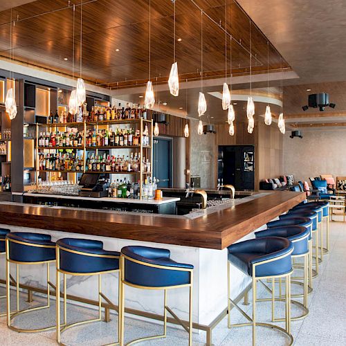 The image shows a modern bar with a wooden countertop, blue chairs, and hanging light fixtures, surrounded by a stocked bar and lounge seating area.