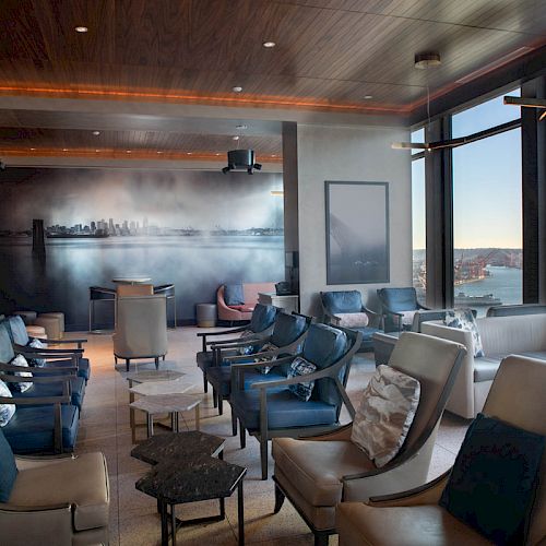 The image shows a modern, upscale lounge area with seating, light fixtures, large windows overlooking water, and a mural of a city skyline on one wall.