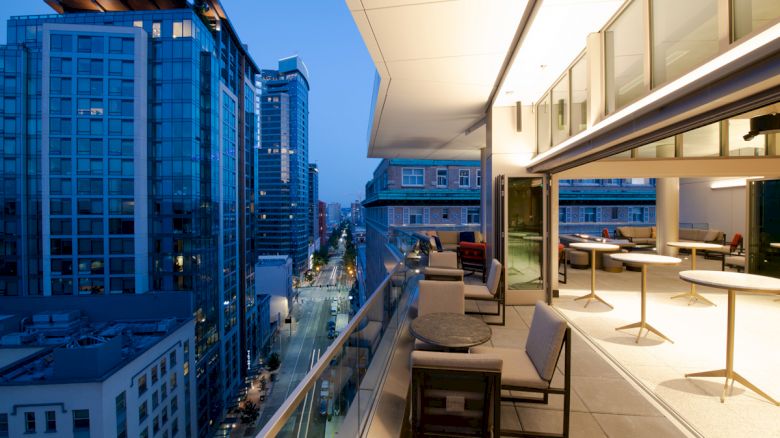 A modern balcony view with outdoor seating overlooks a vibrant cityscape with tall buildings and bustling streets at dusk, offering a serene atmosphere.