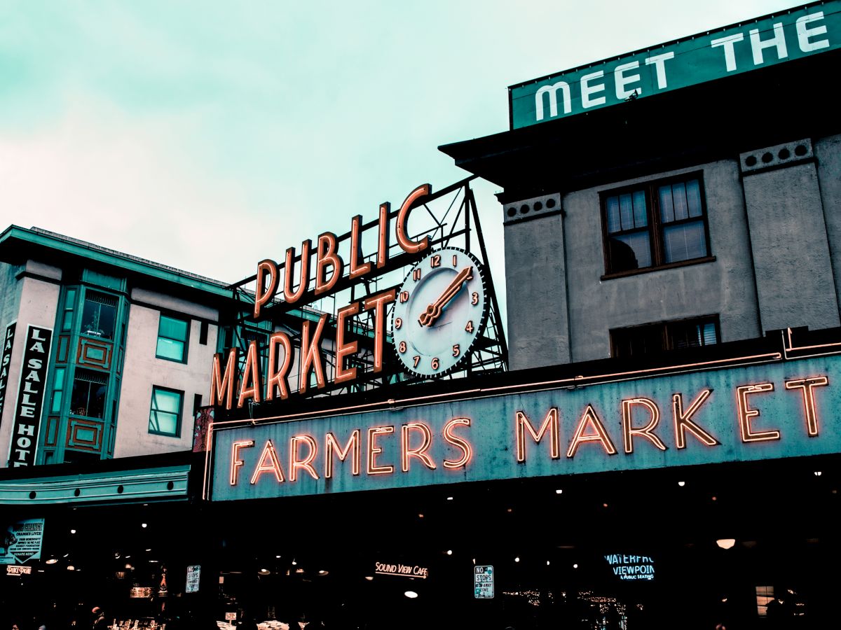 The image shows a public market with a neon sign reading "Farmers Market" and a clock. Buildings and a hotel sign are visible in the background.