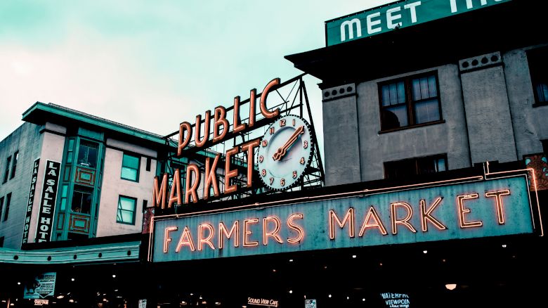 The image shows a public market with a neon sign reading "Farmers Market" and a clock. Buildings and a hotel sign are visible in the background.