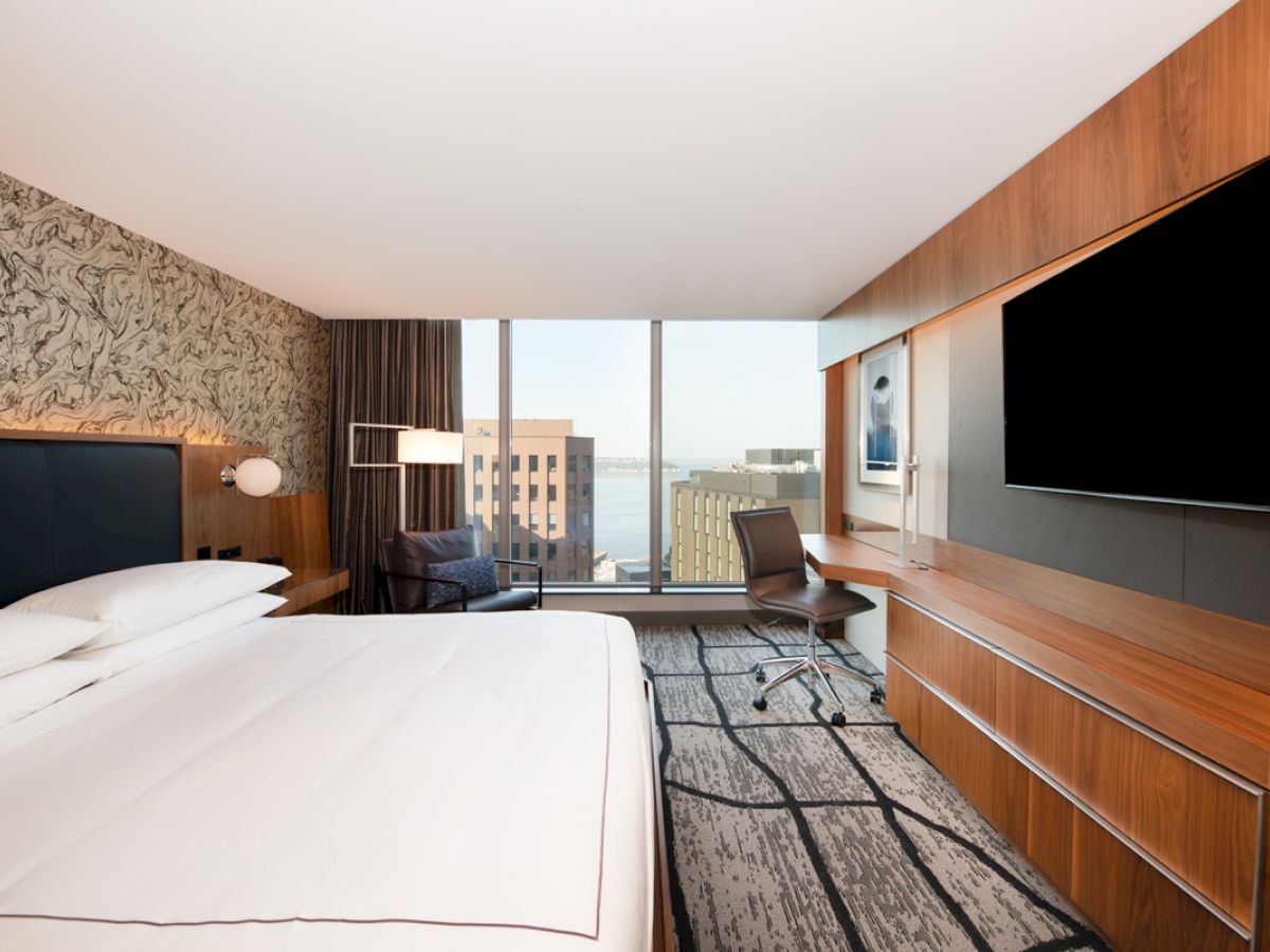 A modern hotel room with a large bed, wall-mounted TV, desk, chair, and a window offering a cityscape view with buildings in the background.