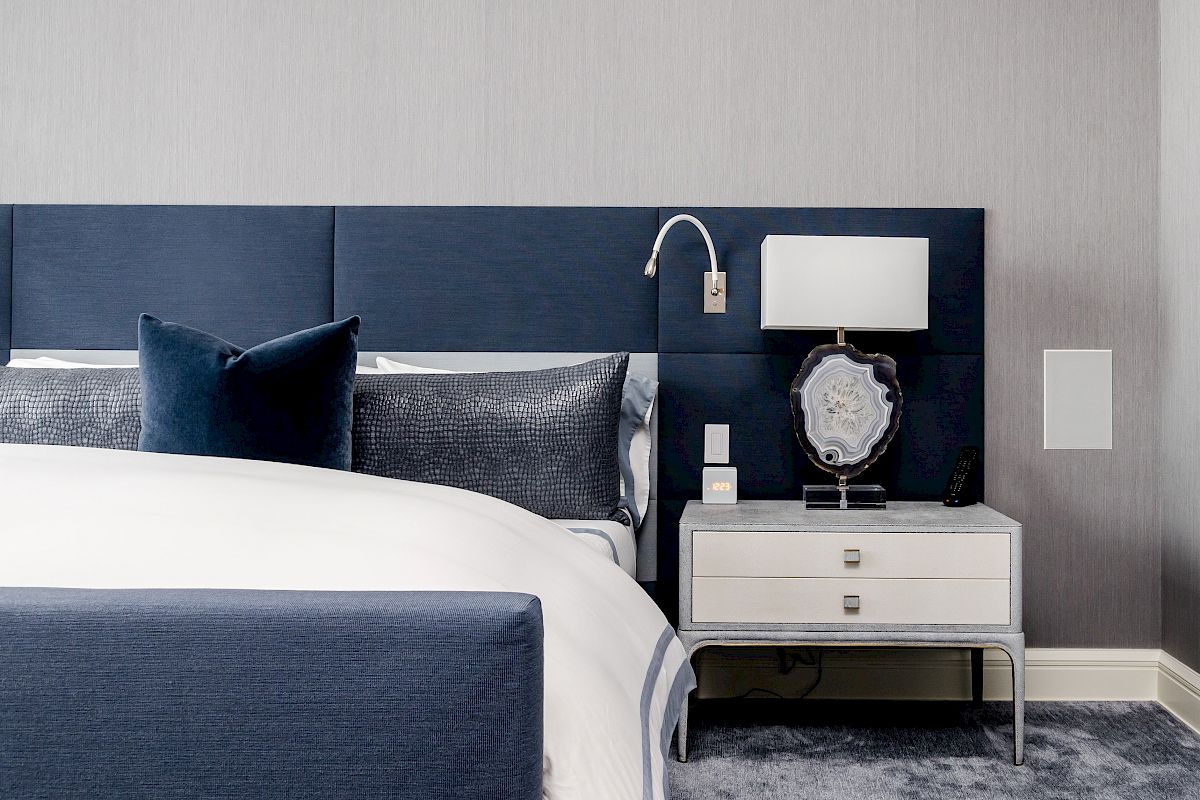 A stylish bedroom featuring a blue upholstered bed, a gray carpet, and a nightstand with a lamp, decorative piece, and a wall-mounted reading light.