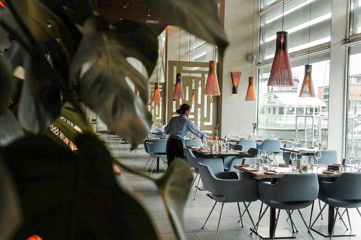 A person is setting tables in a modern restaurant with large windows and stylish lighting.