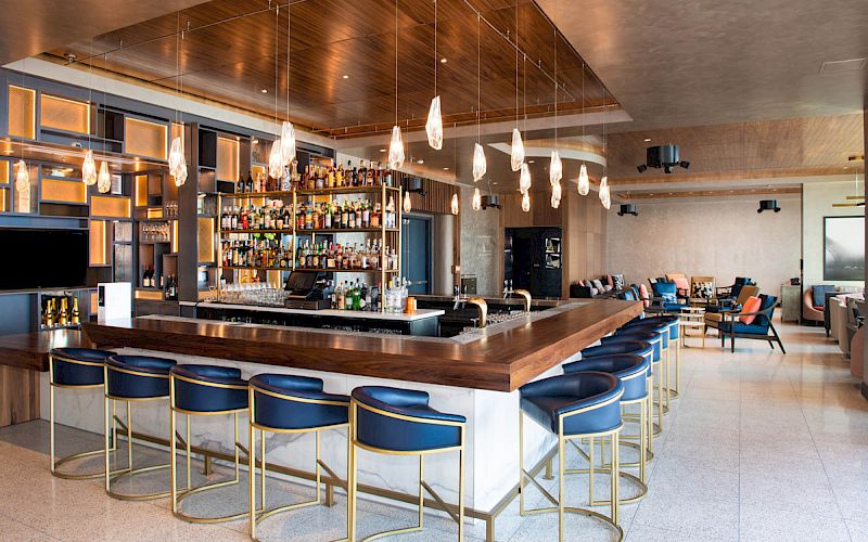 A modern bar with a wooden countertop, blue stools, bottle shelves, pendant lights, and lounge seating in the background.