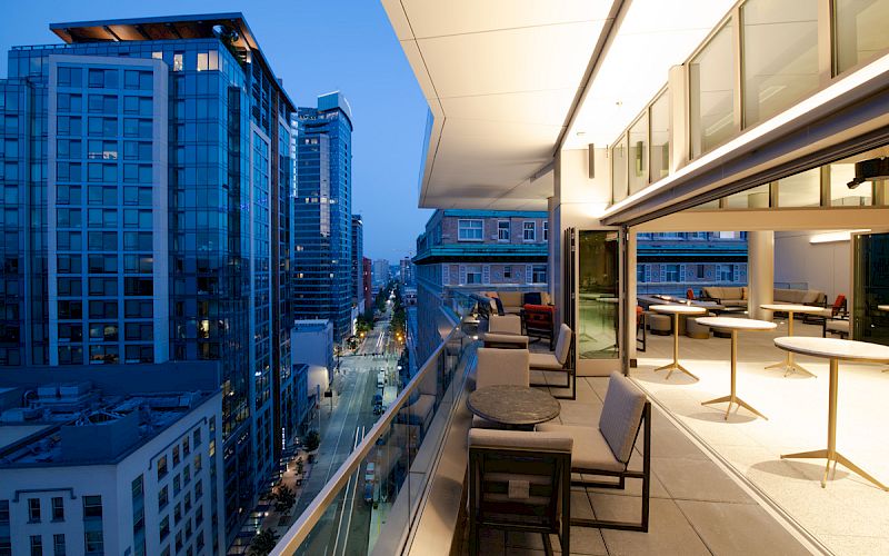 An outdoor balcony with tables and chairs overlooks a city street with modern high-rise buildings at dusk, creating a cozy urban atmosphere.