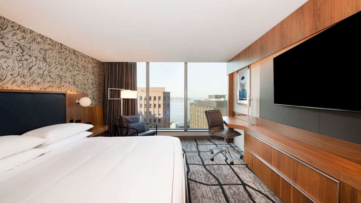A modern hotel room with a large bed, wall-mounted TV, desk, chair, and a window offering a view of city buildings and water in the distance.