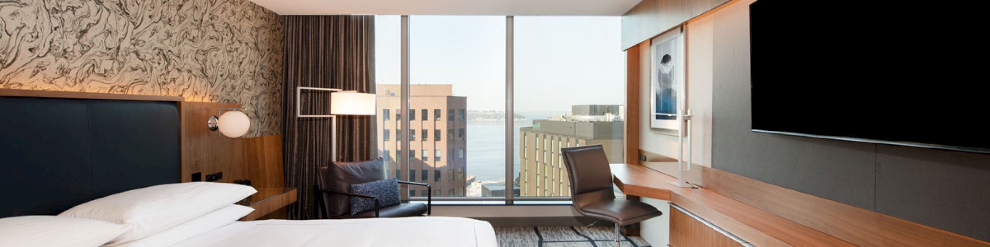 A modern hotel room with a large bed, wall-mounted TV, desk, chair, and a window offering a view of city buildings and water in the distance.